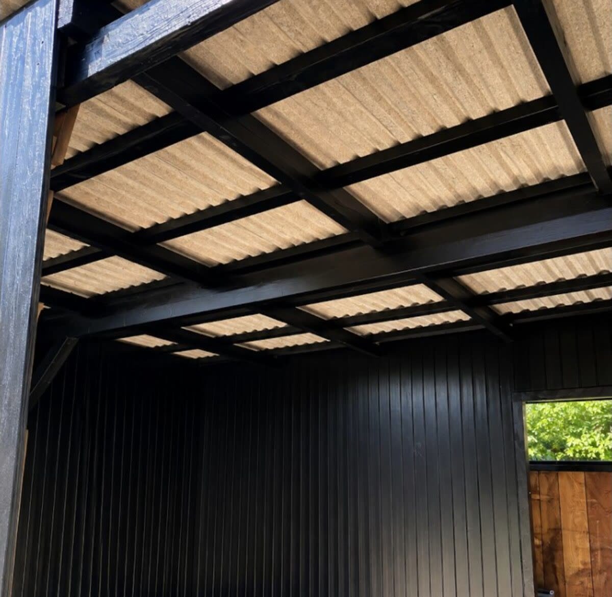 Sortmalet træbeklædning og bjælker under bølgepladetag i carport